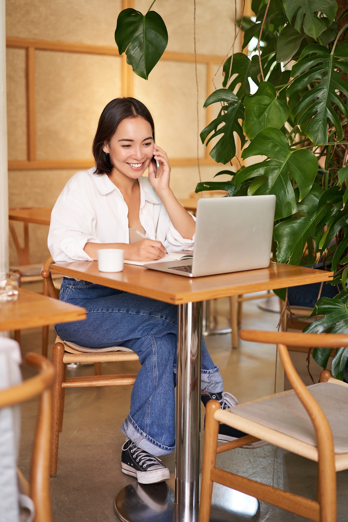 Stylish young woman answer phone call, sitting in cafe with laptop, working and taling on